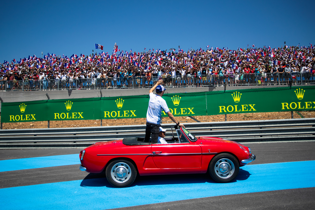 Formula 1 driver Esteban Ocon waves at fans in a grandstand from a vintage car